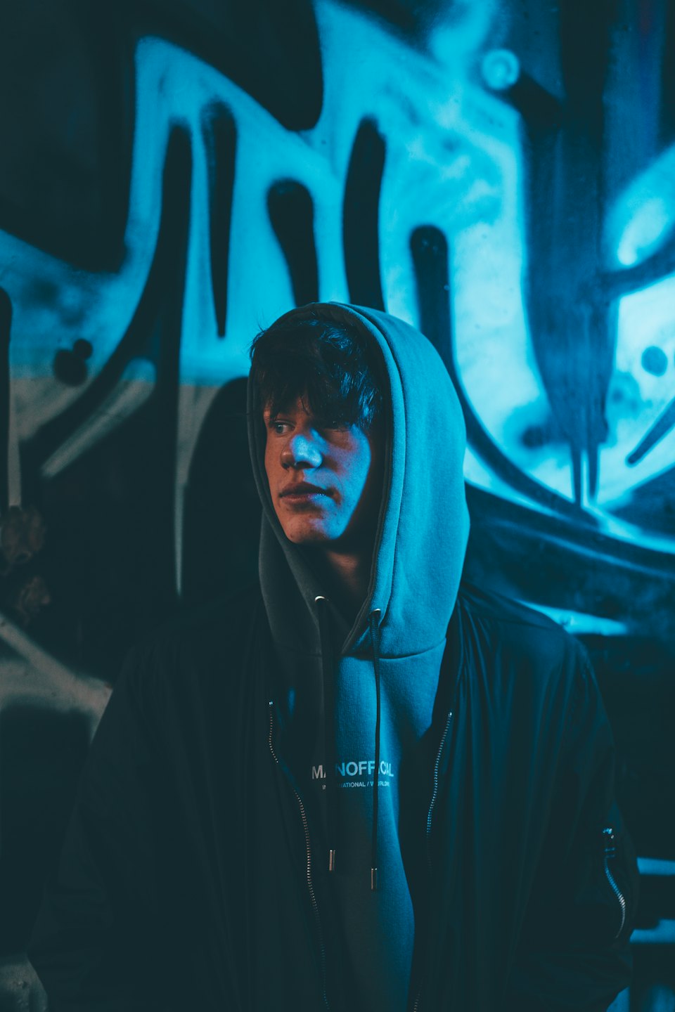 Young man in hoodie illuminated by blue light near graffiti.